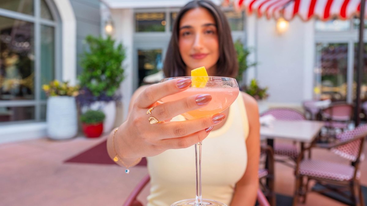 a woman holding out a cocktail toward the camera; she is sitting an an outdoor table near a striped umbrella at Juno & The Peacock
