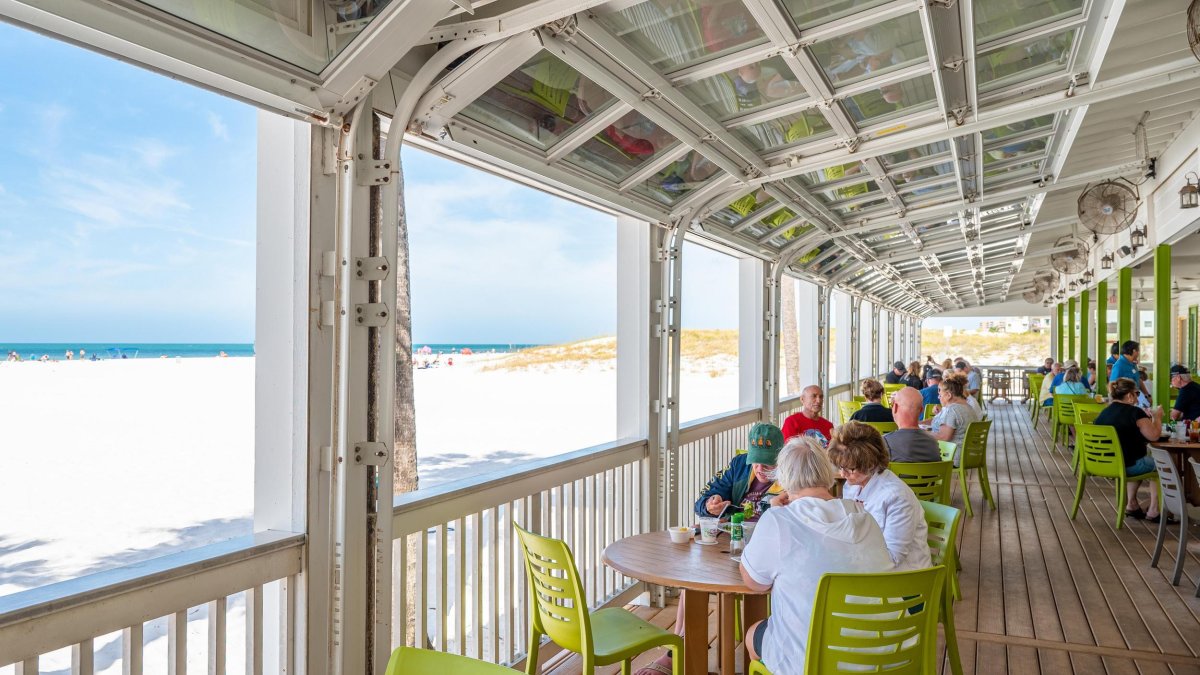 Diners at tables on the deck of Crabby's Beachside Pavilion, with a view of white sand and the Gulf in the distance.