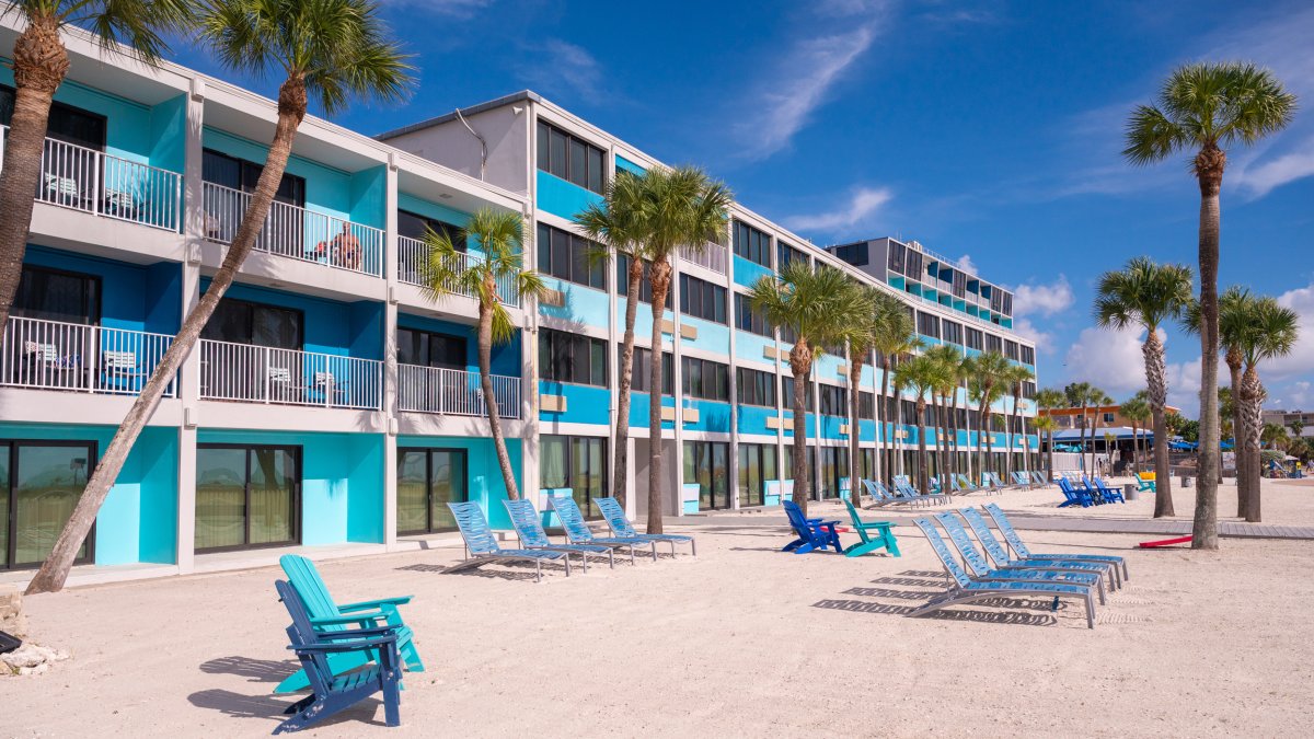Outside of Bilmar Beach Resort's turquoise and white building with tall palm trees outside and beach chairs on the sand in Florida.