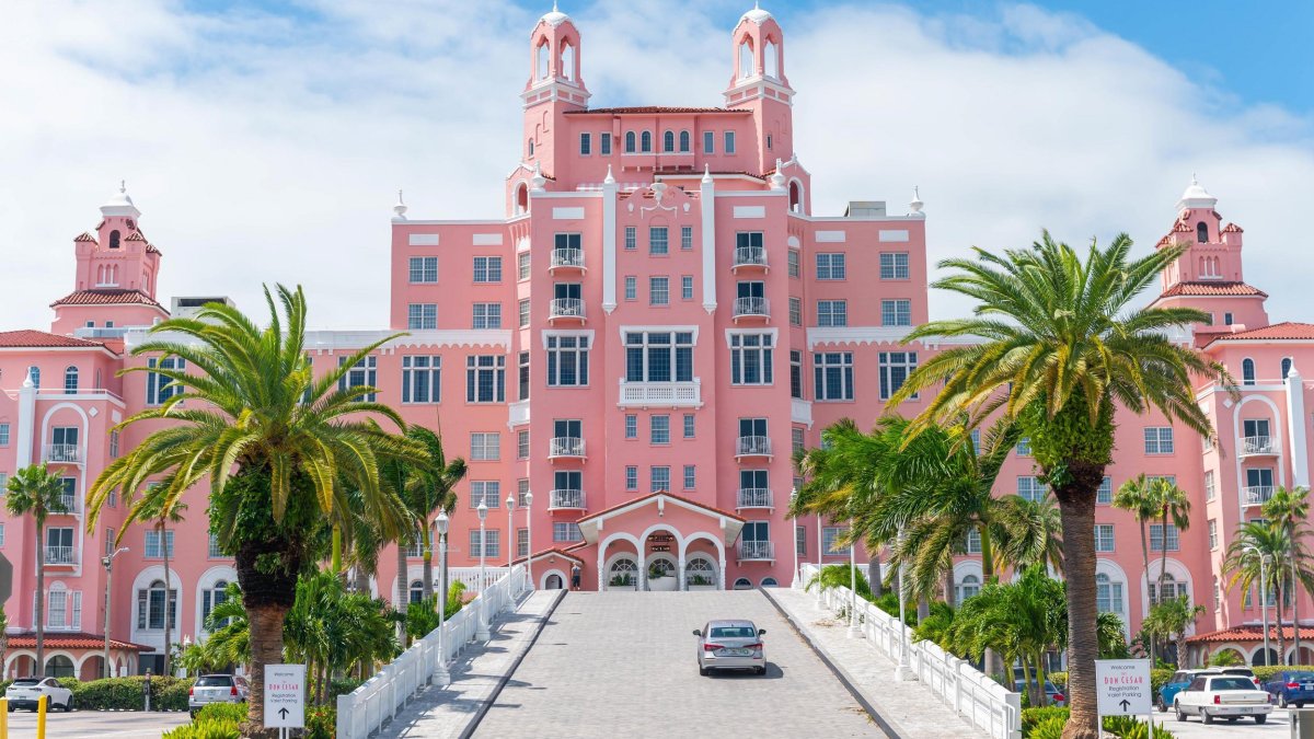 The Don CeSar hotel with bright pink exterior and almost castle-like structure, with palm trees off the side and a silver car driving towards it in Florida.