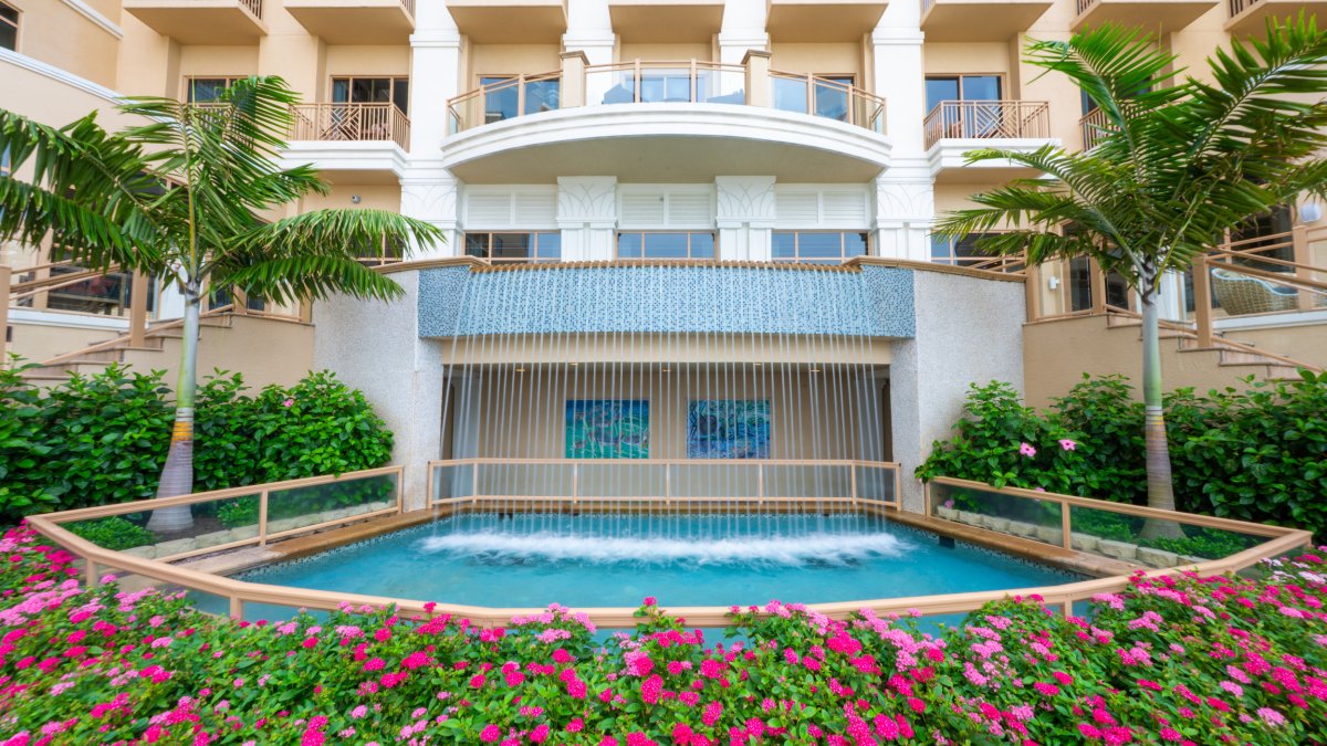 Exterior view of Sandpearl Resort with vibrant pink flowers below and a waterfall with palm trees on each side in Florida.