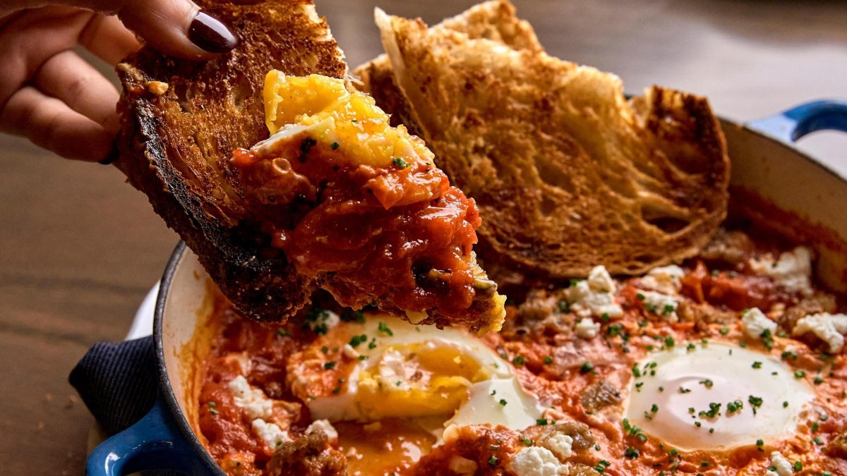 a person's hand dipping toasted bread into a large pan of Shakshouka during brunch at Elliott Aster at The Vinoy Hotel