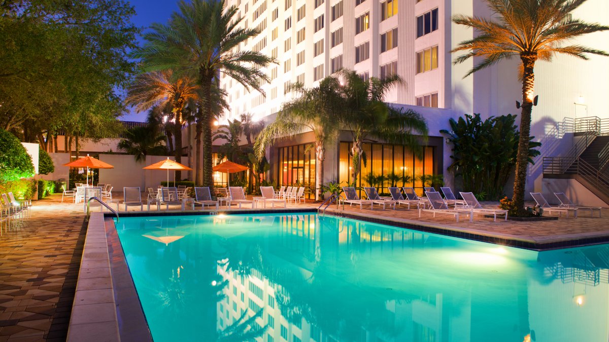Illuminated outdoor hotel pool surrounded by lounge chairs, umbrellas and palm trees at dusk.