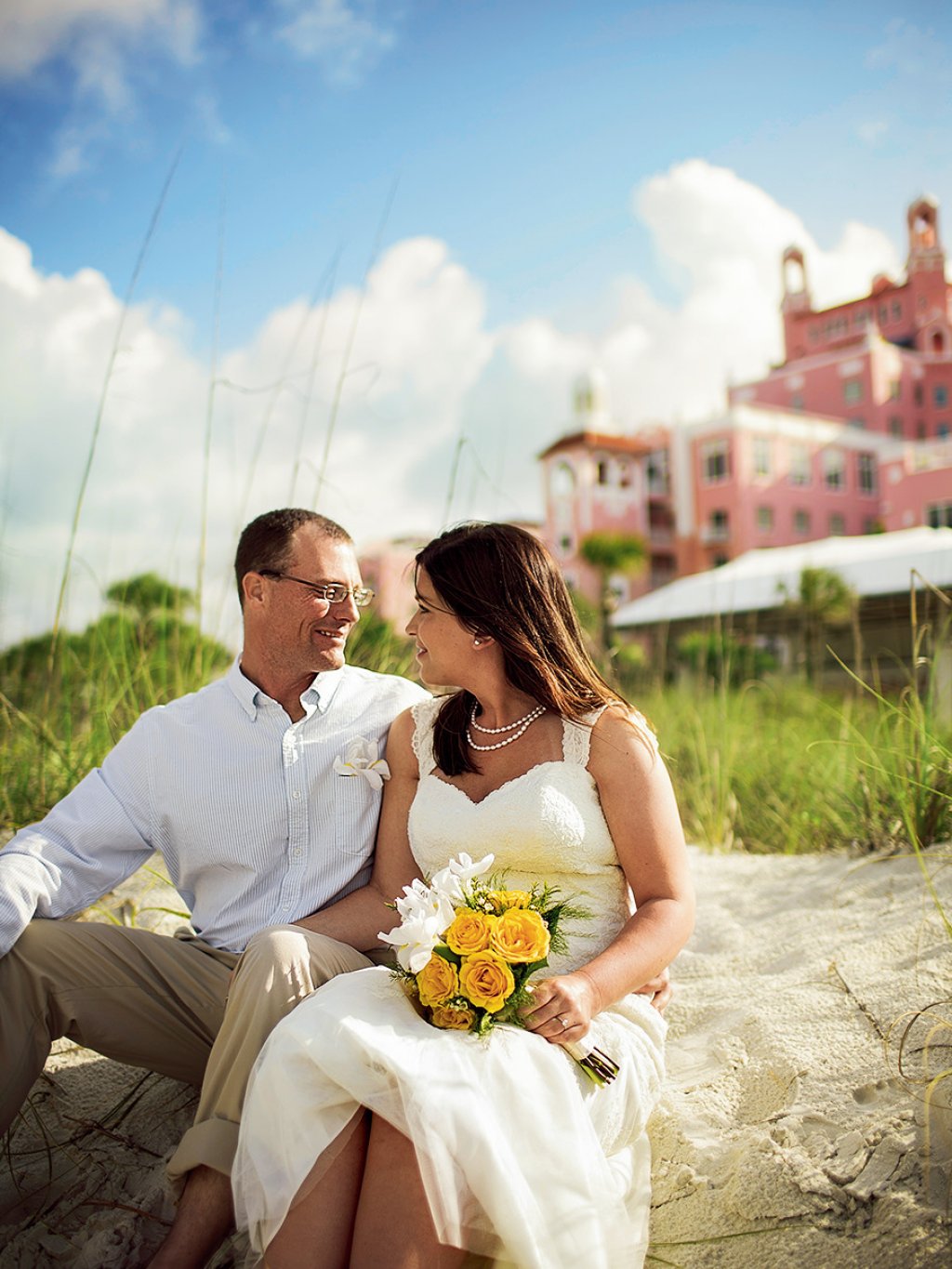 Bride and groom seating on the sand in front of Don Cesar hotel, looking at each other. Bride is holding a bouquet of yellow roses.