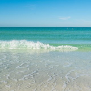 a small wave rolls up on a white beach, the Gulf water is turquoise and clear and the sky is blue