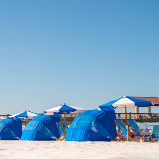bright blue cabanas and blue and white beach umbrellas line a white sand beach with a pier in the background