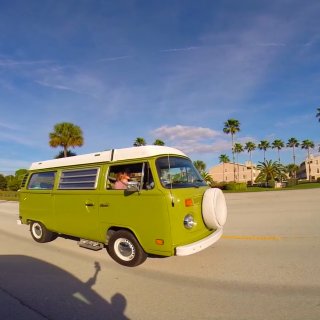 a vintage green van driving on a road with buildings and palm trees in the background