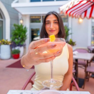 a woman holding out a cocktail toward the camera; she is sitting an an outdoor table near a striped umbrella at Juno & The Peacock
