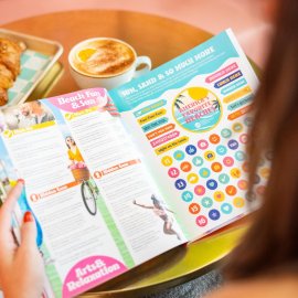 a person holding a magazine designed in bright colors at a small table that has a latte and a buttery croissant on it