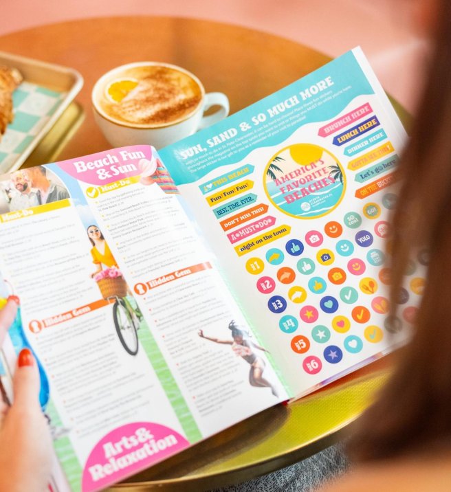 a person holding a magazine designed in bright colors at a small table that has a latte and a buttery croissant on it