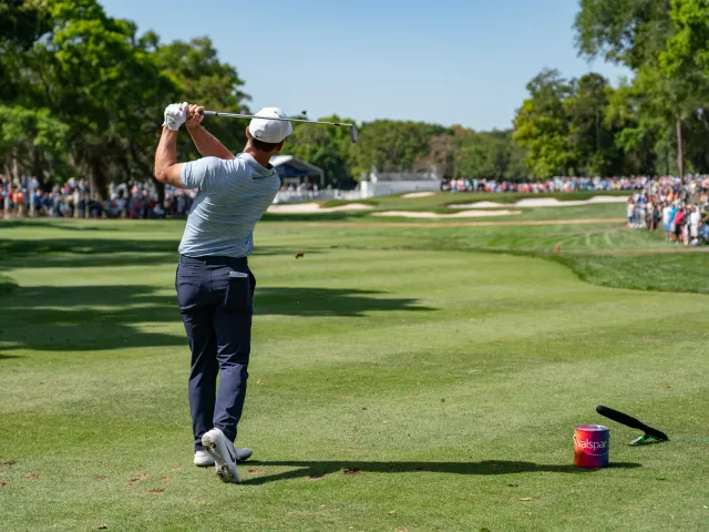 A golfer right after hitting the ball. A large crowd can be seen at a distance.