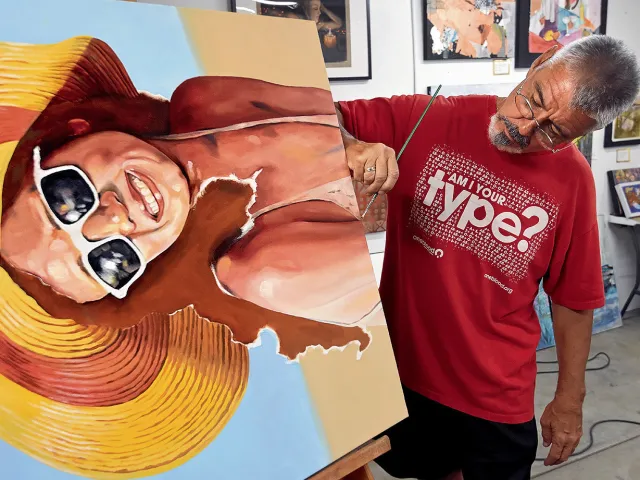 Painter Bob Ribits working at Soft Water Studios in the Warehouse Arts District finishing a painting of a lady in swimsuit, sunglasses and beach hat.