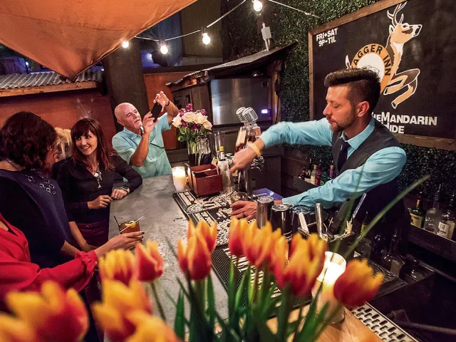Group of people sitting at a bar with a bartender pouring drinks. Foreground has a vase full of lilies