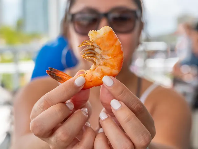 A lady wearing sunglasses holding a shrimp right in front of the camera.