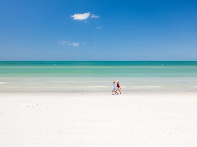 Two beachgoers walk along the white sandy shoreline and emerald green waters of St. Pete Beach on a bright sunny day.