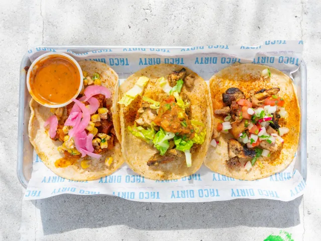 Three different tacos with topping on a horizontal tray with a side of salsa at Taco Dirty in St. Pete