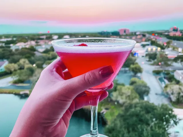 a manicured hand holding a pink cocktail with a cherry with a view of sunset and the Gulf from Vantage Rooftop Bar at the Karol Hotel