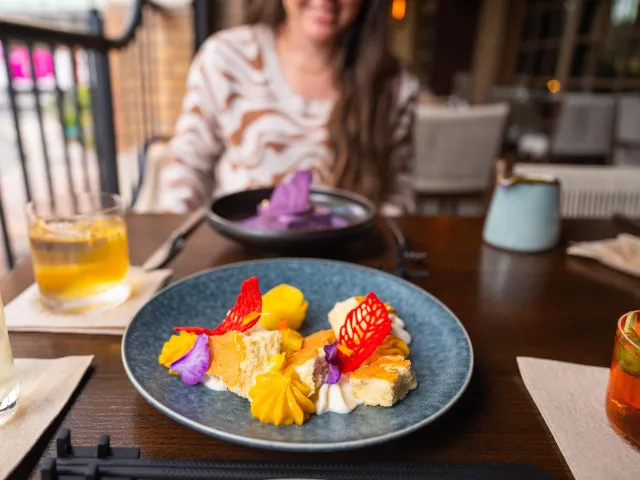 a colorful plate of food with cocktail surrounding it and a young woman in the background at Fortu restaurant