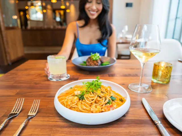 a smiling woman in a bright blue dress sits across from a bowl of pasta at a restaurant
