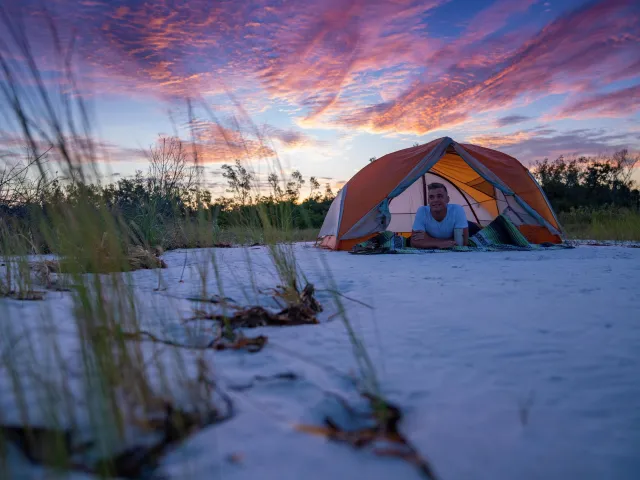 a young man relaxes inside his tent on the beach at Shell key with a dramatic pink and blue sky behind him