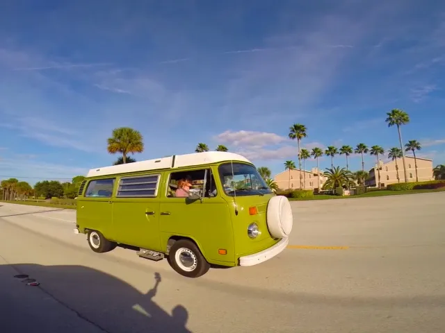 a vintage green van driving on a road with buildings and palm trees in the background