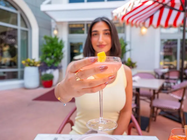 a woman holding out a cocktail toward the camera; she is sitting an an outdoor table near a striped umbrella at Juno & The Peacock