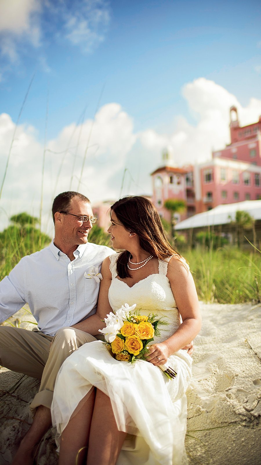 Bride and groom seating on the sand in front of Don Cesar hotel, looking at each other. Bride is holding a bouquet of yellow roses.