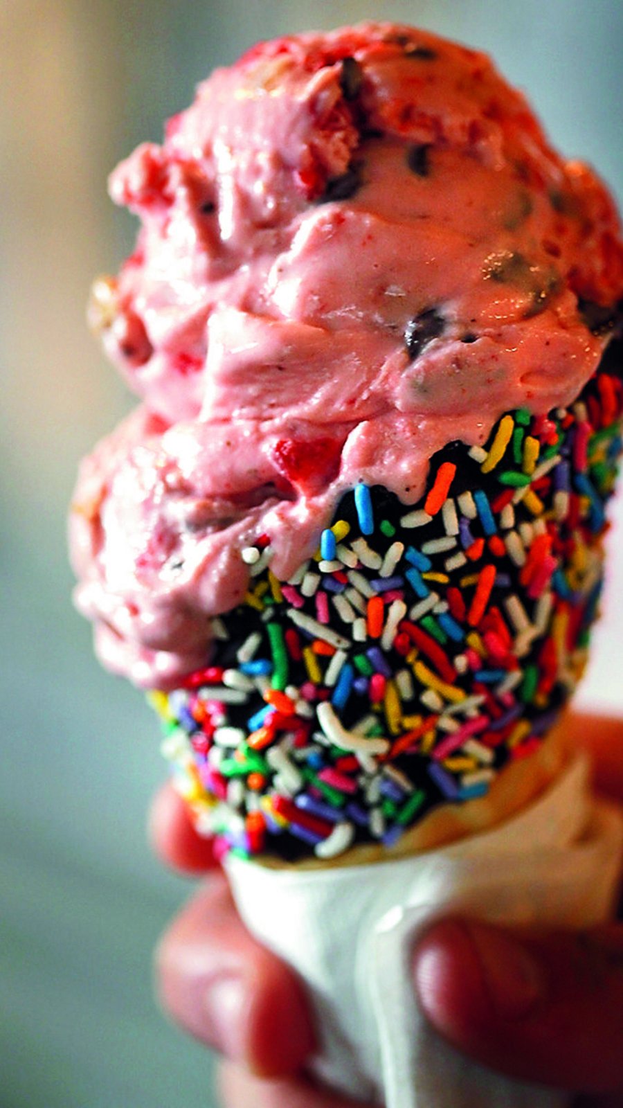 hand holding a pink ice cream cone on a waffle cone covered in chocolate and rainbow sprinkles.