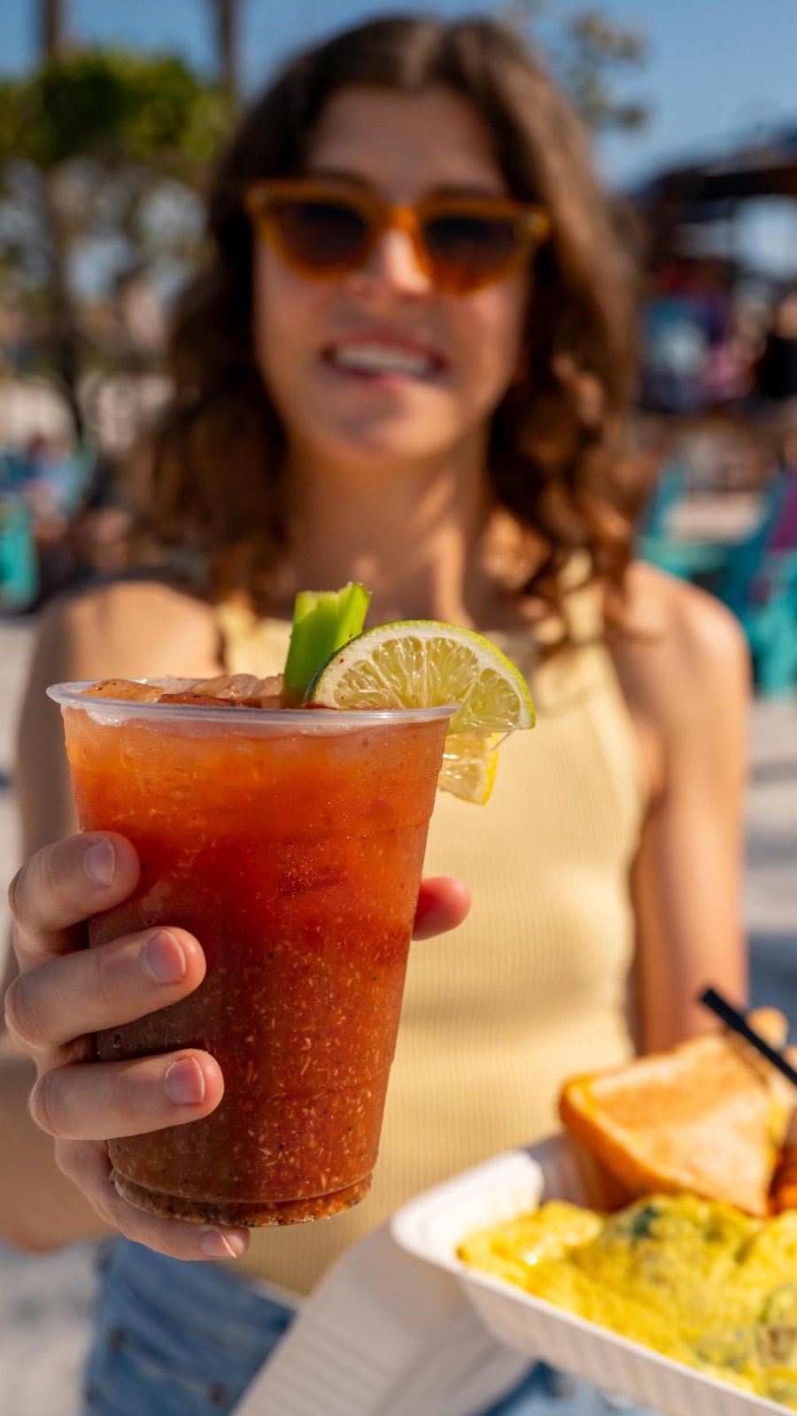 A woman holds a cocktail and breakfast dish on the beach