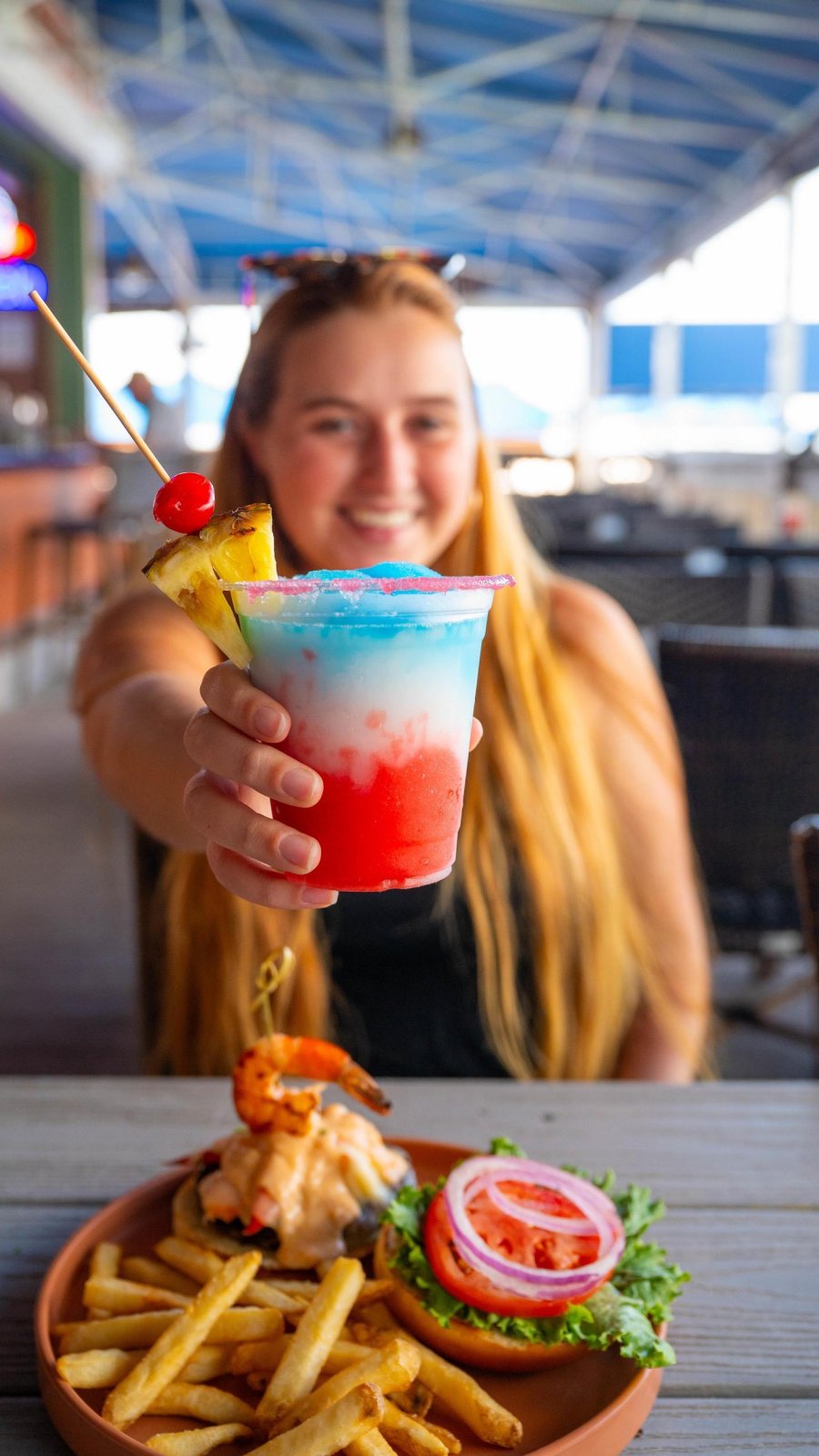 A young woman holds up a festive drink at a beachside restaurant