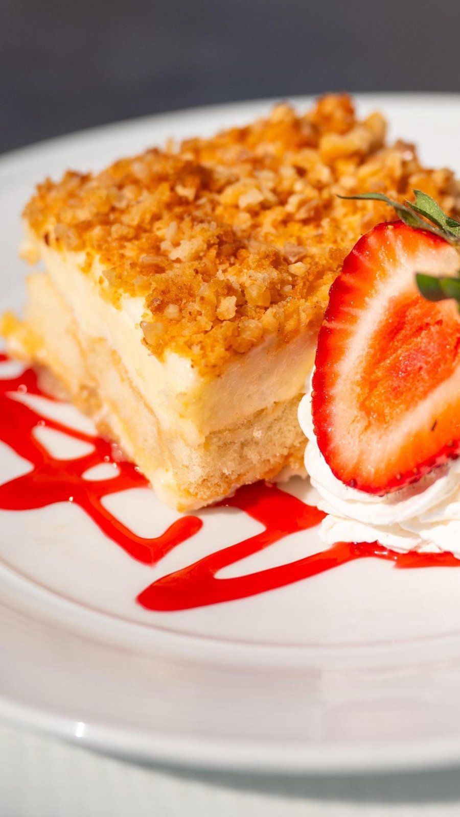a square cheesecake slice with a crumble top, garnished with whipped cream and strawberries and a drizzle of strawberry sauce