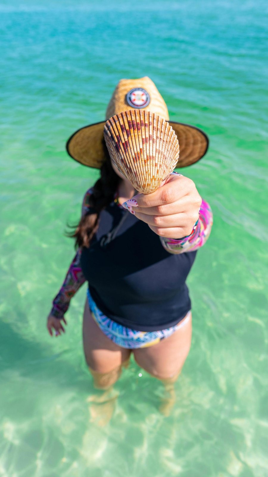 a soman standing in clear, green, knee-deep water and holding a giant cockle shell in her hand