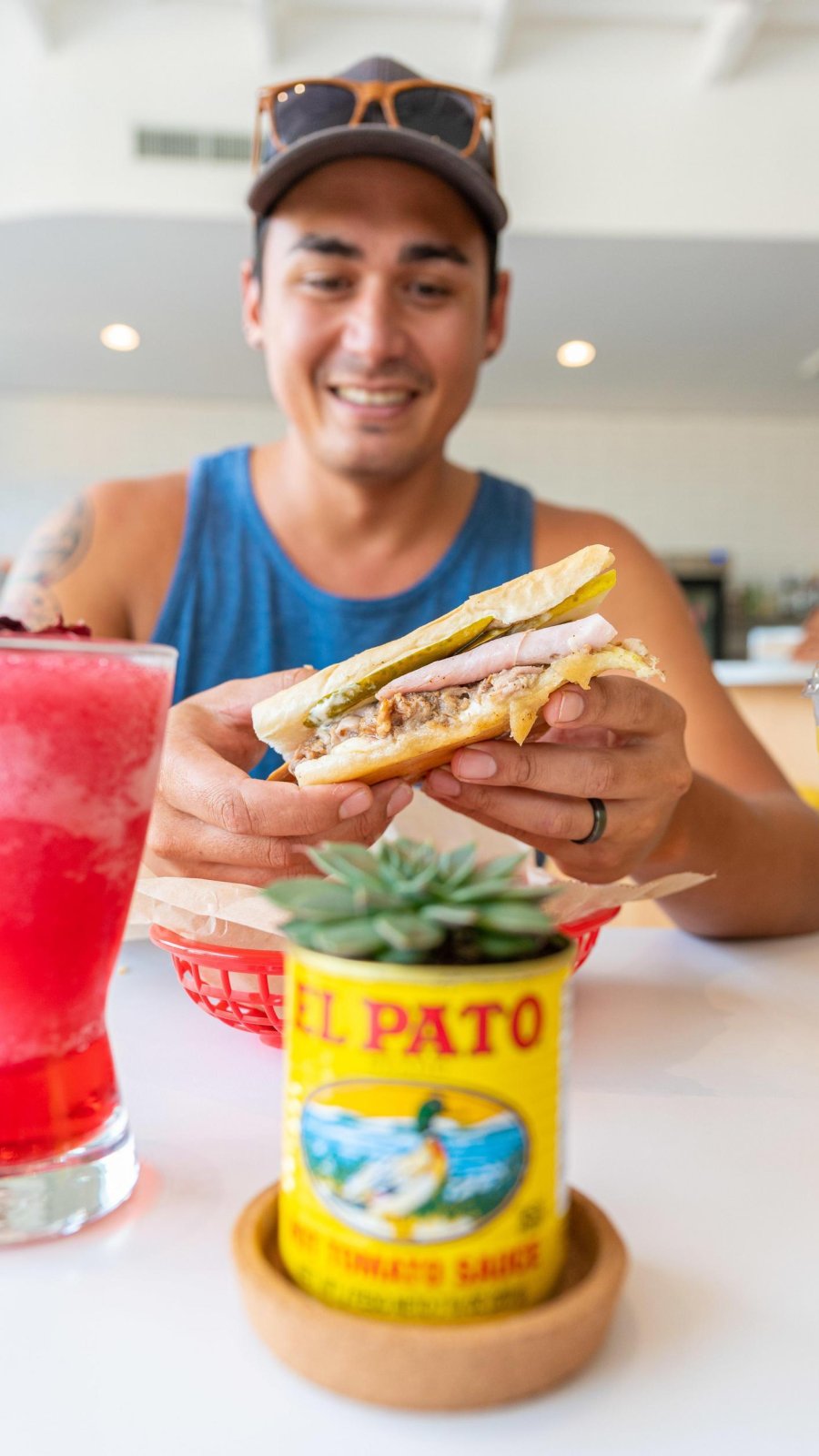 a man holds a Cuban sandwich, with a bright pink frozen drink in front of him at a table at Bodega in St. Pete