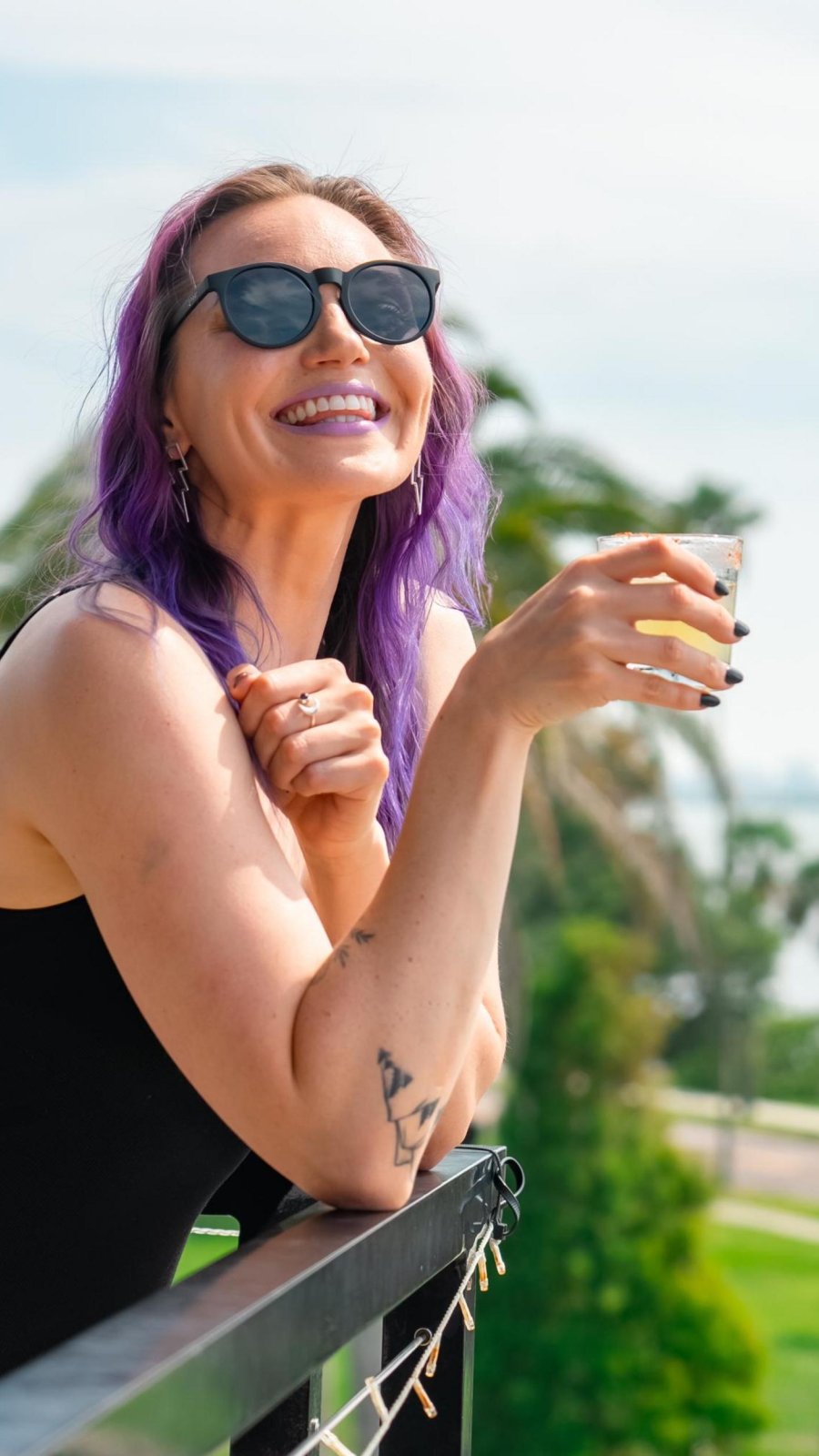 a smiling woman who is holding a cocktail rests her arm on deck of the Hi-Fi Rooftop Bar at the Fenway Hotel in Dunedin