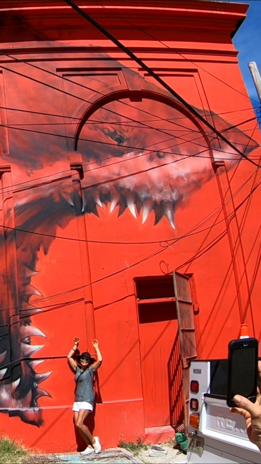a woman poses, pretending to be frightened, next to a bright red mural of a great white shark on a building in St. Pete