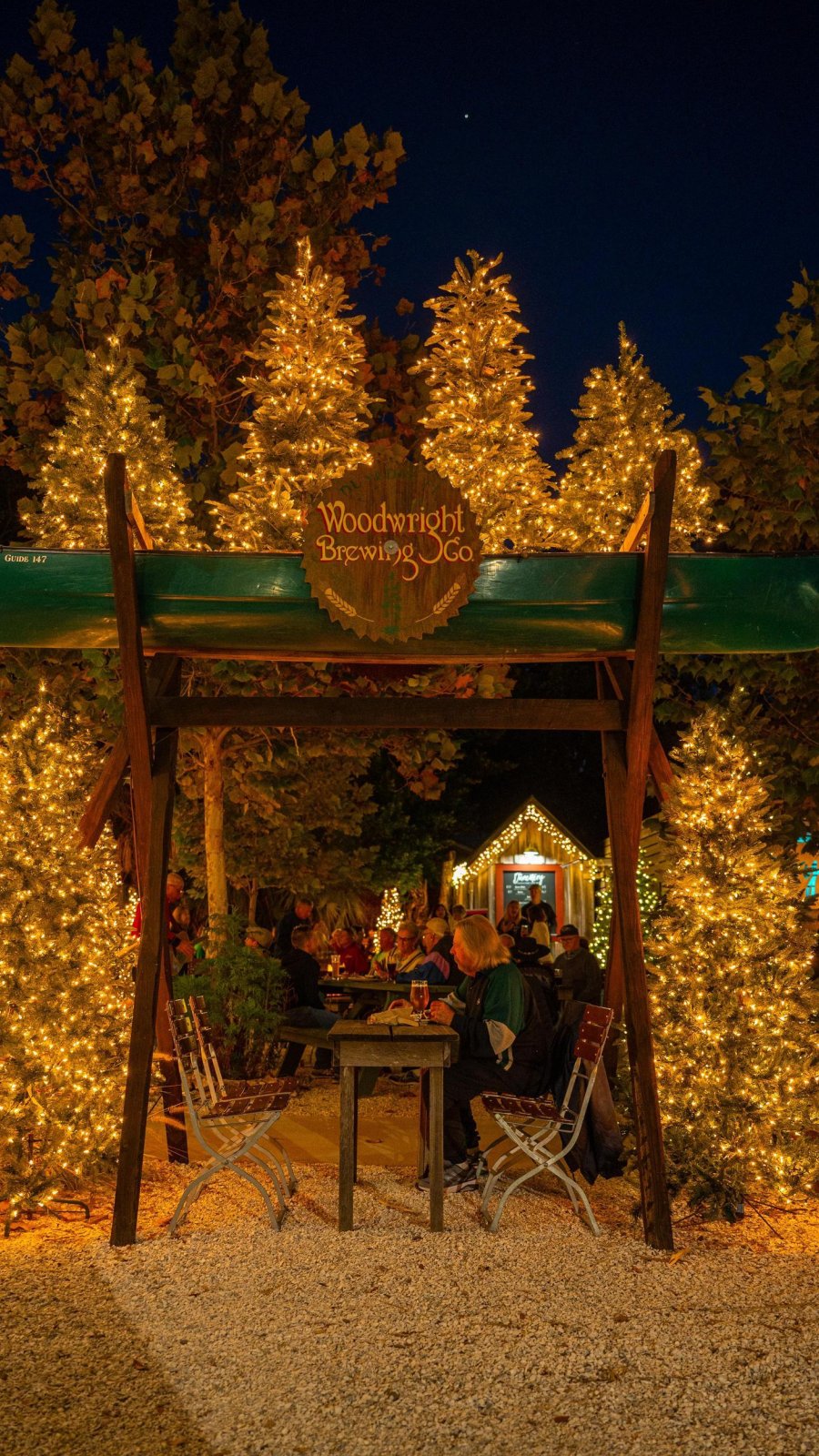 people drinking beer outside under a shelter surrounded by holiday lights and Christmas trees at Woodwright Brewing Co.