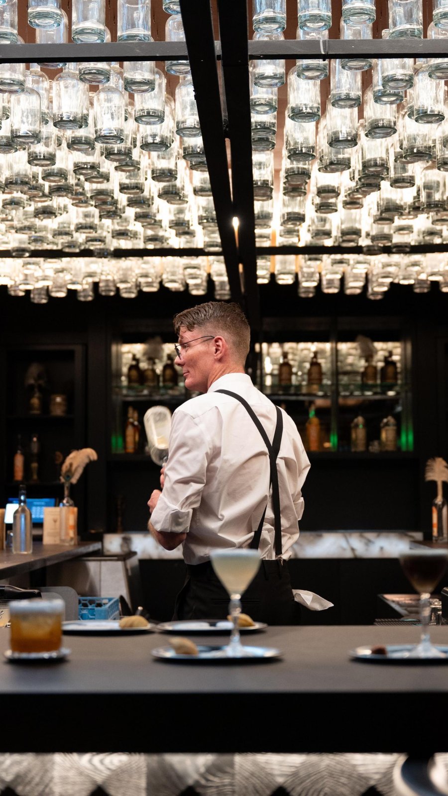 Bartender behind the bar at 31 South Speakeasy beneath a ceiling installation of illuminated glass bottles.